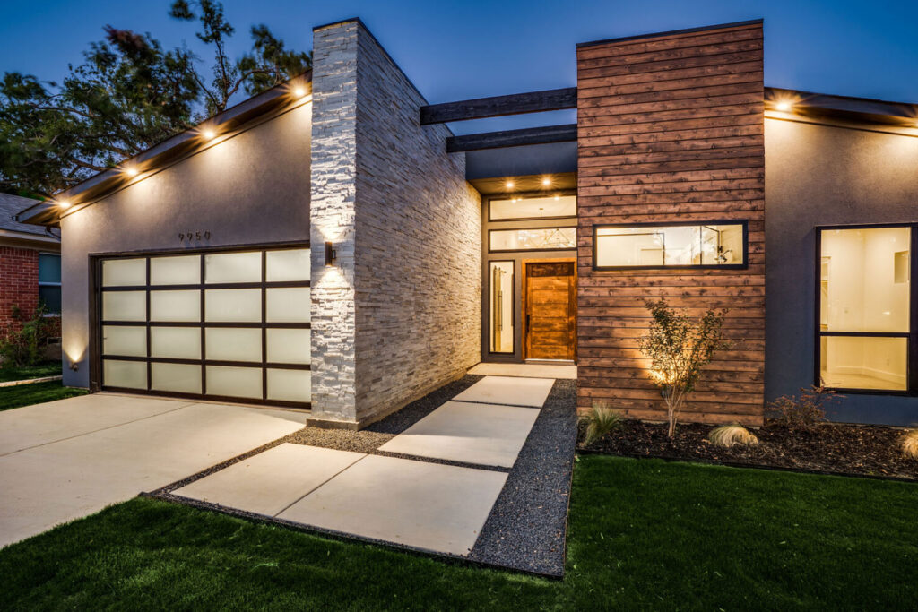 Modern home entry with a stacked stone column beside the garage, wood cladding and landscape lighting